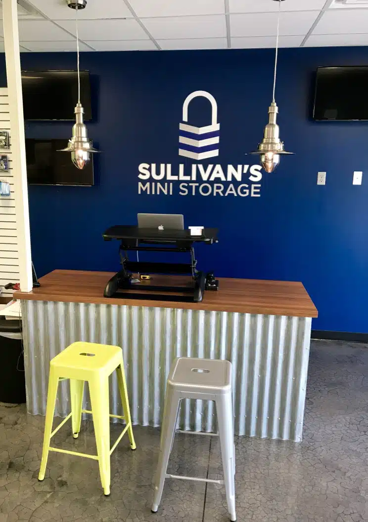 Reception area of Sullivan’s Mini Storage with a blue accent wall, logo, front desk, and two metal stools.