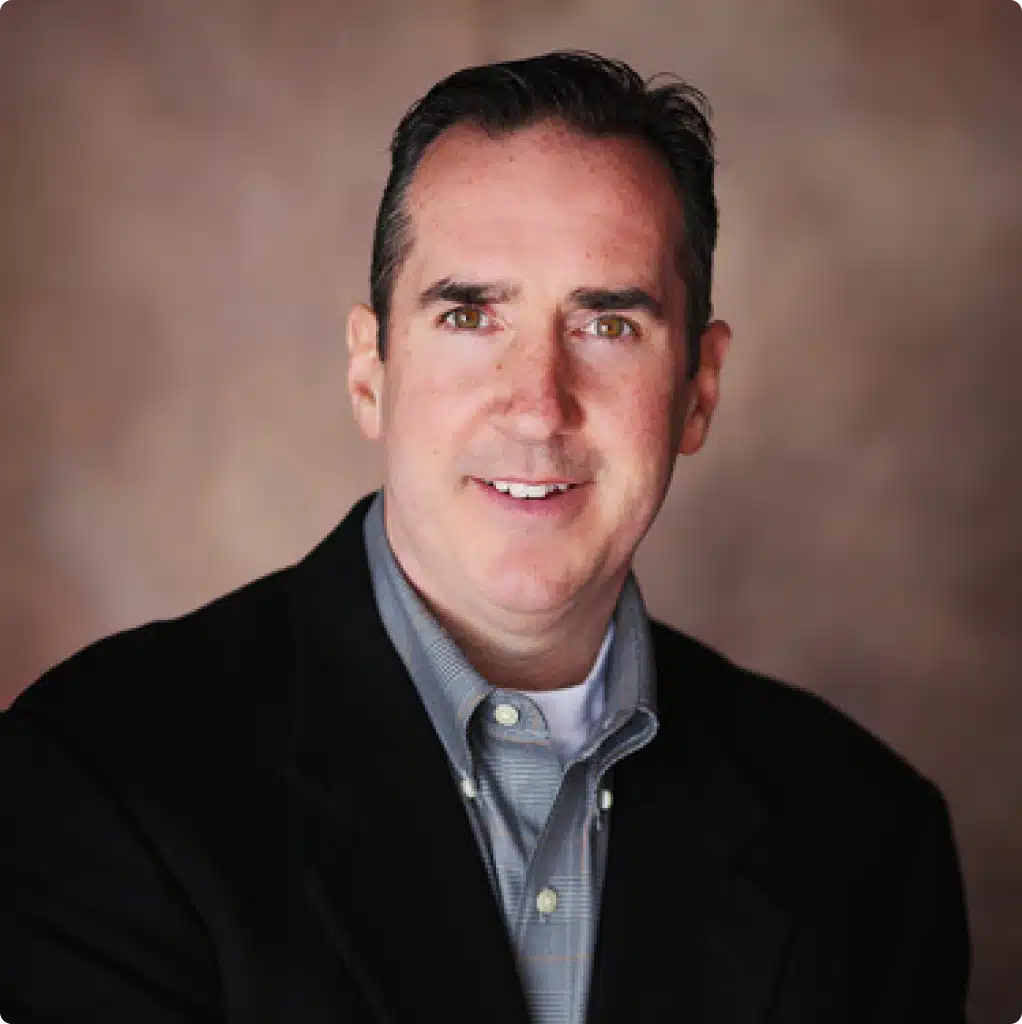 Photograph of a smiling man with brown hair and eyes, wearing a black blazer and gray button-down shirt, Mike Sullivan